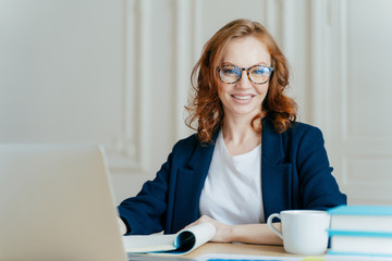 Wall Mural - Glad smiling red haired young female concentrated on creating new business project, owns corporation, sits in front of laptop computer, wears optical glasses and elegant outfit, updates software