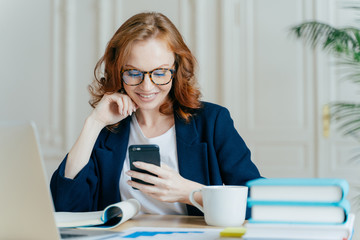 Wall Mural - Photo of lovely female sits with smartphone device, types feedback, works in office on up to date laptop, focused into screen of gadget, sits at work place with books, notepad and hot beverage