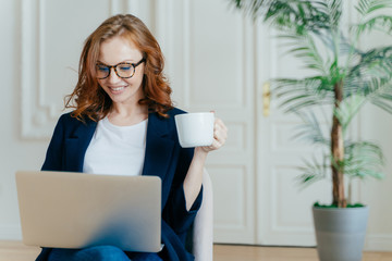 Wall Mural - Optimistic red haired young successful female freelancer works remotely in coworking space, makes research, does online courses, sits in armchair, keyboards on laptop computer, holds cup of hot drink.