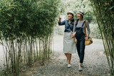 Fototapeta Bambus - full length asian women having fun together outdoors on hike through amazing bamboo forest trail. young girl pointing finger on tree showing sharing talking with friend. cheerful female tourist kyoto