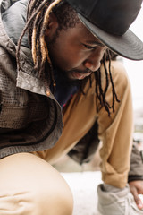 Wall Mural - Handsome African American with dreadlocks in brown trousers and a blue sweater in a black cap on his head against the background of houses, city