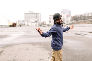 Wall Mural - Handsome African American with dreadlocks in brown trousers and a blue sweater in a black cap on his head against the background of houses, city