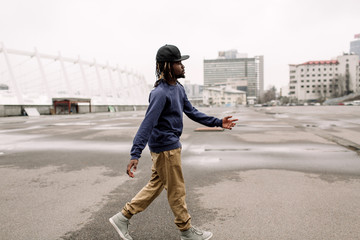 Wall Mural - Handsome African American with dreadlocks in brown trousers and a blue sweater in a black cap on his head against the background of houses, city