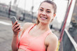 © Artem - Portrait of happy young sports woman holding smart phone in hand and listening to music in earphones while standing on bridge. Attractive fitness girl smiles and listens motivational playlist on phone