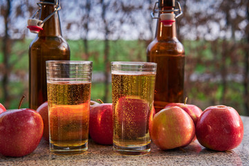 Close up Picture on two glasses or jars full of organic sparkling apple cider and two rustic bottles with red riped apples on stone garnit table in the garden restaurant during summer sunny evening.