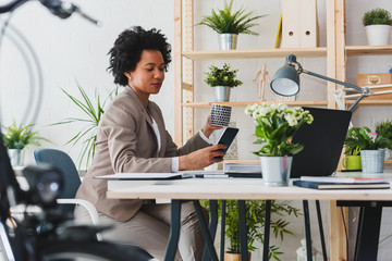 Wall Mural - Happy smiling african-american business woman working on laptop at office, using smart phone. Businesswoman sitting at her working place