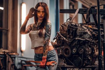 Female car mechanic holding a big hammer and posing next to a car engine suspended on a hydraulic hoist in the workshop.