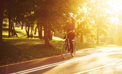 Young attractive woman riding through the park after work. Beautiful lady cycling during sunset. Bike as a trendy transport. Healthy outdoors activity on a warm summer day. Bicycle trend in the city.