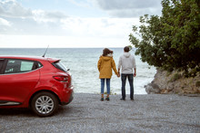 Rear View Of Young Tourist Couple Standing On Rocks Near His Car On A Winter Holiday With Coats, Looking At The Sea. Travel Lifestyle Exterior.