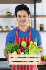 Close up portrait of happy young Asian handsome bearded man wearing blue t shirt and striped apron hold wooden box full of vegetables, presenting healthy meals images in modern kitchen background