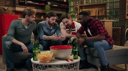 Four young male friends using smartphone, man in the center shows the screen of the phone, the rest of the company dismiss him and leave the room, bearded attractive young man sits alone surprised