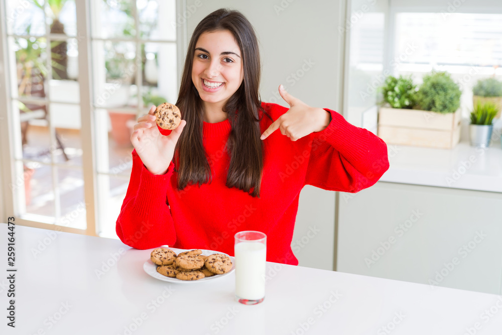Beautiful young woman eating chocolate chips cookies and glass of milk with surprise face pointing finger to himself
