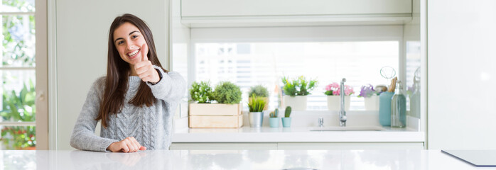 Poster - Wide angle picture of beautiful young woman sitting on white table at home doing happy thumbs up gesture with hand. Approving expression looking at the camera with showing success.