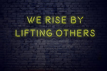 Positive Inspiring Quote On Neon Sign Against Brick Wall We Rise By Lifting Others