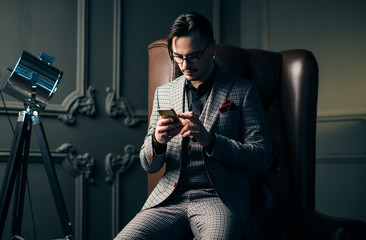 Wall Mural - Young handsome man posing for a fashion shoot in a studio. Fashion as a lifestyle. Man wearing a beard. Model sitting on a sofa in tuxedo. Successful fashionable businessman. Business look advertising