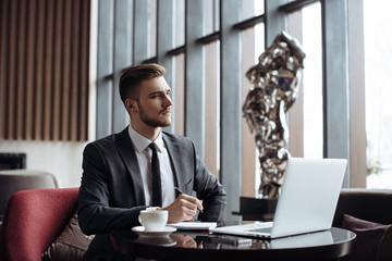 Wall Mural - Young handsome man sitting in office with cup of coffee and working on project connected with modern cyber technologies. Businessman with notebook trying to keep deadline in digital marketing sphere.