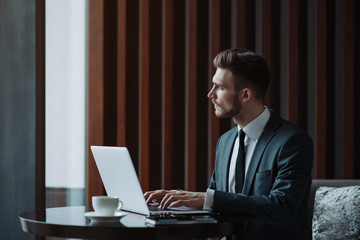 Wall Mural - Young handsome man sitting in office with cup of coffee and working on project connected with modern cyber technologies. Businessman with notebook trying to keep deadline in digital marketing sphere.