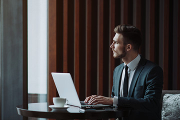 Wall Mural - Young handsome man sitting in office with cup of coffee and working on project connected with modern cyber technologies. Businessman with notebook trying to keep deadline in digital marketing sphere.