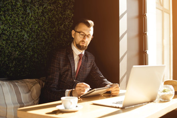 Young handsome man sitting in office with cup of coffee and working on project connected with modern cyber technologies. Businessman with notebook trying to keep deadline in digital marketing sphere.