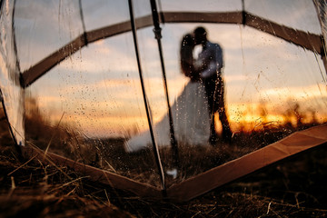 the bride and groom in the rain are covered with a transparent umbrella, rain drops. Sunset.