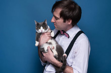 Handsome Man In Shirt, Suspender And Pink Bow Tie Holding And Looking To Cute Grey Cat With White Paws On Blue Background With Copy Space