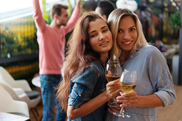  Portrait of two attractive women posing with glasses of wine at party in bar, their male friends dancing in background