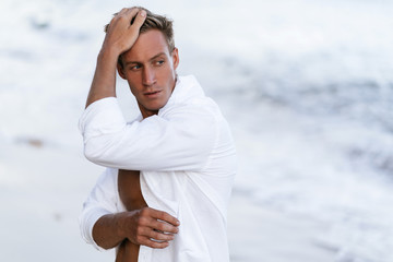 Wall Mural - Portrait of handsome sexy man in white shirt posing on beach, ocean waves at background