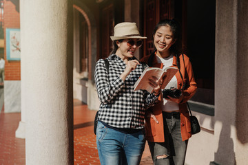 two happy cheerful smiling asian girl travelers walking in corridor at Confucian temple. young female tourists holding camera guide book and talking chatting about history information on sunset time.