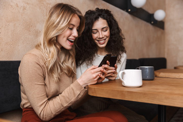 Wall Mural - Emotional shocked girls friends sitting in cafe using mobile phone.