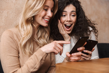 Wall Mural - Emotional shocked girls friends sitting in cafe using mobile phone.