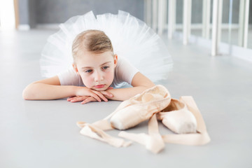 Wall Mural - Little ballerina is looking at pointe shoes in ballet class. Cute child girl is dreaming and lying on floor. Tired kid is wearing white dress with tutu skirt after dance training.