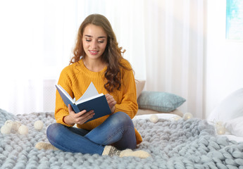 Canvas Print - Young beautiful woman in warm sweater reading book on bed at home, space for text