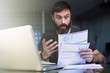 © KleverLevel - Bearded positive man working with laptop at home browsing documents . Businessman going through paperwork at home office with unpaind bills.