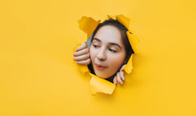 Funny Teenage Girl Peeping Through Hole On Yellow Paper. The Concept Of Surprise, Joyful Mood From What He Saw. Discounts, Sales, Surprise. Copy Space.