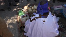 High Angle View Of Rear Side Of Black African Man, Traditional Healer With His Medicines Sitting Low On The Pillow On The Pavement Surrounded By Bystanders And Talking To Them