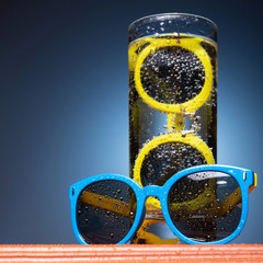 Wall Mural - the concept of summer and leisure, in a glass of water yellow sunglasses, blue sunglasses lie nearby, water drops