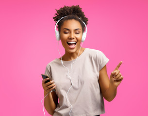 Happy young girl listens music in headphones. Photo of african american girl wears casual outfit on pink background. Emotions and pleasant feelings concept.