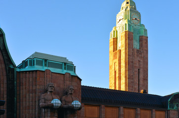 Wall Mural - Helsinki, Finland. View Of Statues On Entrance To Helsinki Central Railway Station in shadows and sun Illuminated clock tower in back ground.