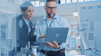 Poster - Female Chief Engineer Talks with Male Electronics Specialist, Explaining Things, He Holds on Laptop Computer. Modern and Bright Office with Stylish People and Working Robot Arm in Background