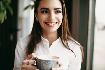 Wall Mural - Close up portrait of a amazing female freelancer looking away smiling while holding a cup of coffee while working in a coffee shop.