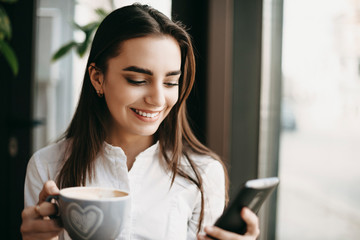 Wall Mural - Attractive caucasian woman using a smartphone smiling while holding a cup of coffee near a window in a coffee shop.