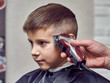 Barber making a haircut to a cute European boy using cutting machine.  Side view.