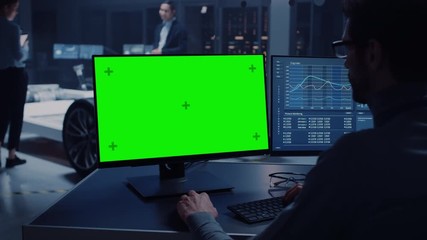 Poster - Professional Engineer Works on a Computer with Green Screen Mock Up and Tests the Electric Car Chassis Prototype with Wheels, Batteries and Engine Standing in a High Tech Development Laboratory.