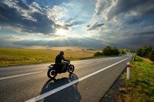Motorcycle Driving On The Asphalt Road In Rural Landscape At Sunset With Dramatic Clouds