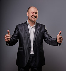 Fun comic bald business man in black suit showing the finger success thumb up sign on grey background. Closeup