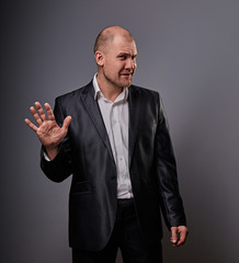 Unhappy depressed bald busuness man in black suit showing the palm stop sign on grey background. Closeup