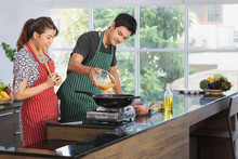 Shot Of Happy Young And Beautiful Asian Couple Sharing Good Times Preparing Lunch Together, Husband Pouring Beaten Eggs Into Hot Pan For Making Omelet In Modern Kitchen Space With Large Glass Window