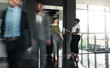 © JonoErasmus - Black african business woman standing with paperwork, waiting to enter a busy elevator lift. With people walking out in a blur, showing motion.