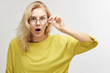 Fototapeta  - Surprised woman in round glasses with open mouth and bulging eyes looks into camera and sees something incredible and amazing isolated on white background