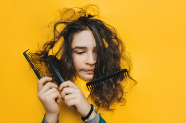 A funny teenage sad girl with problematic curly hair expresses displeasure on her face. Three combs tangled in hard hair.